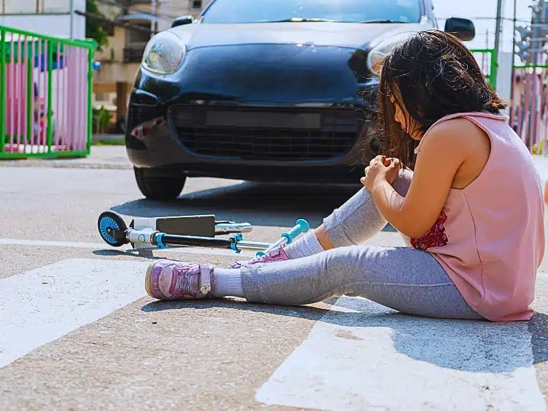 A young child sitting on a crosswalk, appearing hurt, with a fallen scooter nearby. Hit by a Car Outside a Crosswalk