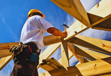 Construction worker hammering wooden roof beams under blue sky.