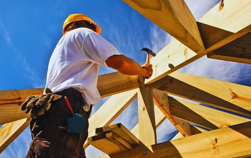 Construction worker hammering wooden roof beams under blue sky.