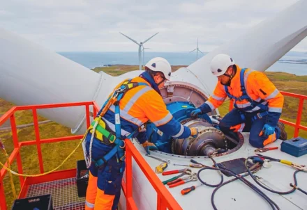 Two technicians in safety gear perform maintenance on a wind turbine nacelle overlooking a coastal wind farm.