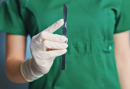 A gloved hand holds a silver scalpel against green scrubs.