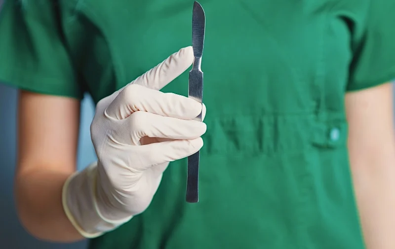 A gloved hand holds a silver scalpel against green scrubs.