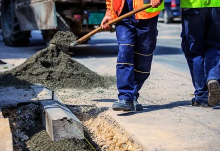 Construction workers in high-visibility vests use shovels to fill a roadside trench with gravel for curb installation.