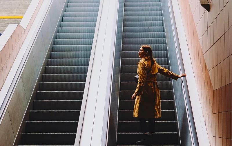 Woman in a tan trench coat standing on an escalator.