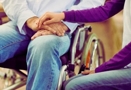 A hand rests supportively on another person in a wheelchair.